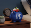 Blue piggy bank labeled 'Dad's Toy Fund' with toys on top, placed on a wooden surface.